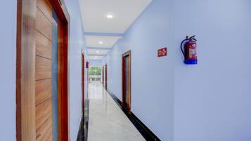 Een hotelgang met een brandblusser aan de muur, een bord met daarop een vluchtroute, en een houten deur aan de linkerkant.