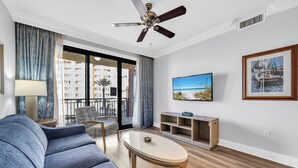 Uma sala de estar moderna com sofá, mesa de centro, TV e varanda com vista para edifícios e palmeiras.