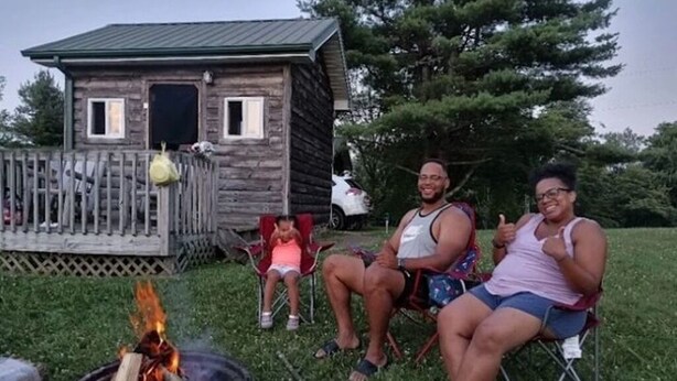 Une cabane rustique avec un foyer au premier plan, entourée d’une terrasse en bois et d’une zone herbeuse.