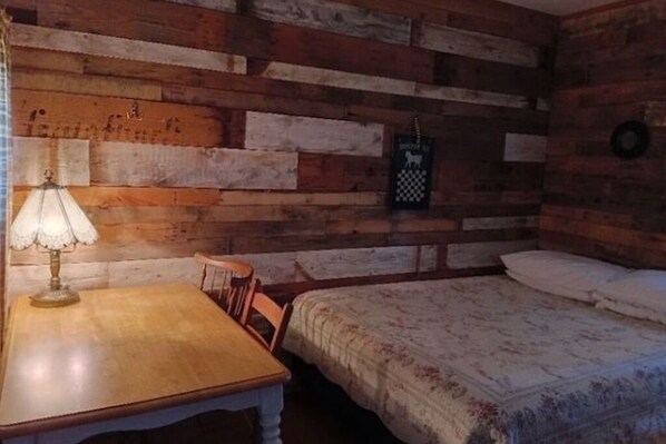A rustic room with a wooden wall, a bed with a floral bedspread, a wooden table with a lamp, and a chair. - Couple's Getaway Camping Cabin with 5-Bathroom Bath House Nearby, Pets Welcome (Fancy Gap)
