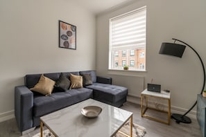En moderne stue med en grå sofa, et hvidt sofabord, en gulvlampe og et vindue med persienner.