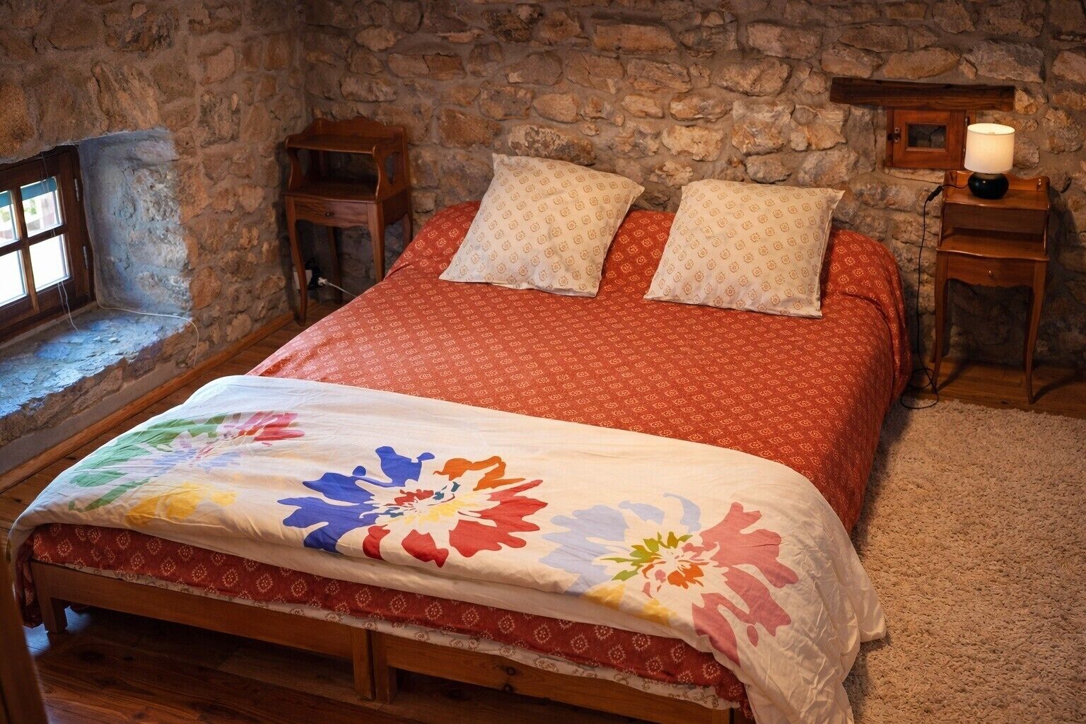 Ein Schlafzimmer mit Steinwand, einem hölzernen Bett, einem Fenster und einem Nachttisch mit Lampe.
