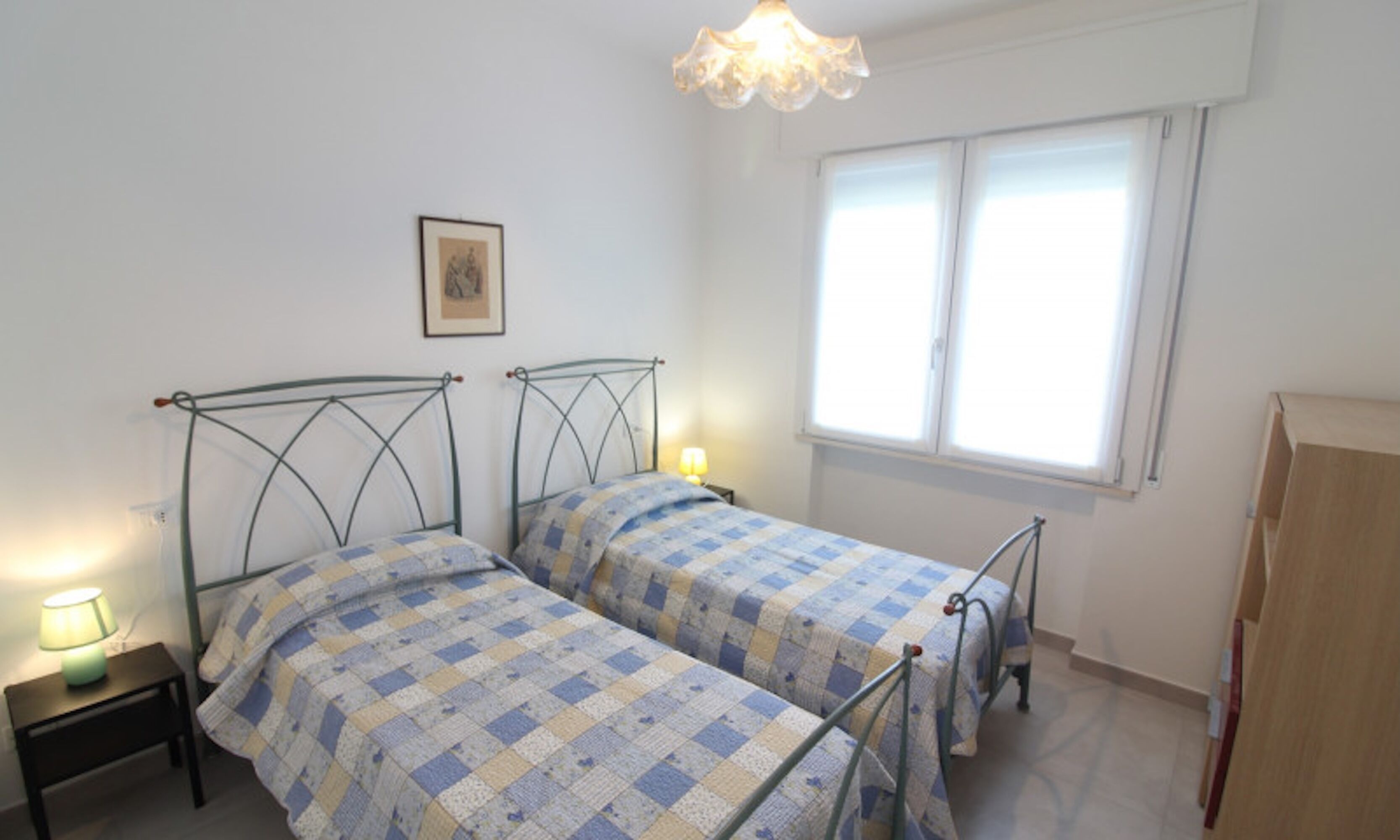 Une chambre avec deux lits, une table de chevet avec une lampe, une fenêtre avec des stores et un tableau encadré au mur.