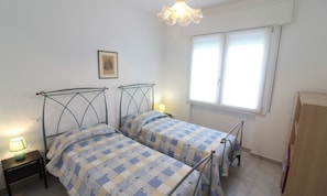 A bedroom with two beds, a nightstand with a lamp, a window with blinds, and a framed picture on the wall. - Coastal Duplex in Lignano Riviera (Lignano Sabbiadoro)