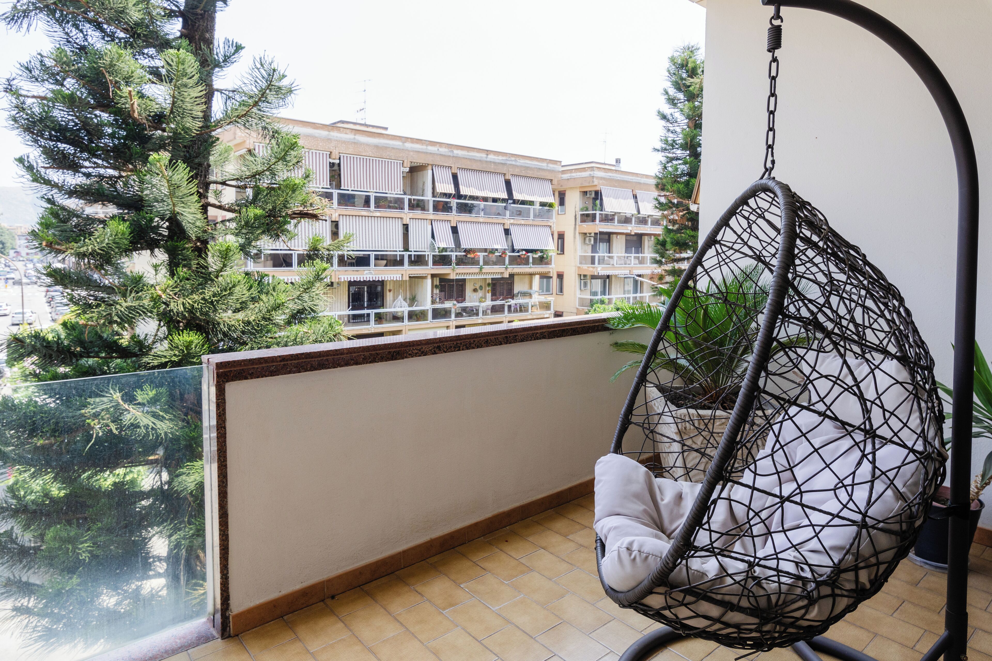Een balkon met een hangstoel, een glazen reling en uitzicht op een gebouw met meerdere balkons.
