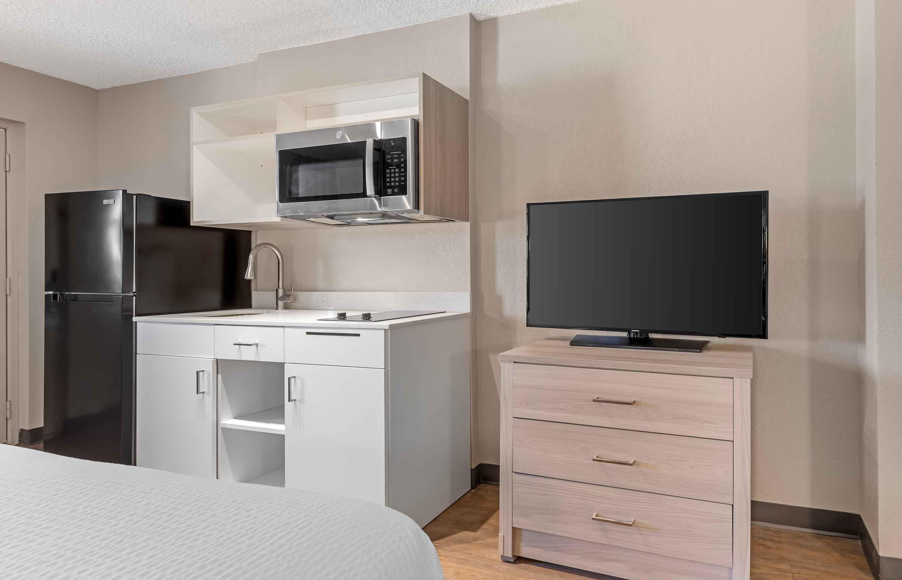 Ein kompaktes Hotelzimmer mit Küchenzeile, einem Flachbildfernseher, einem Bett und einer kleinen Holzkombination.