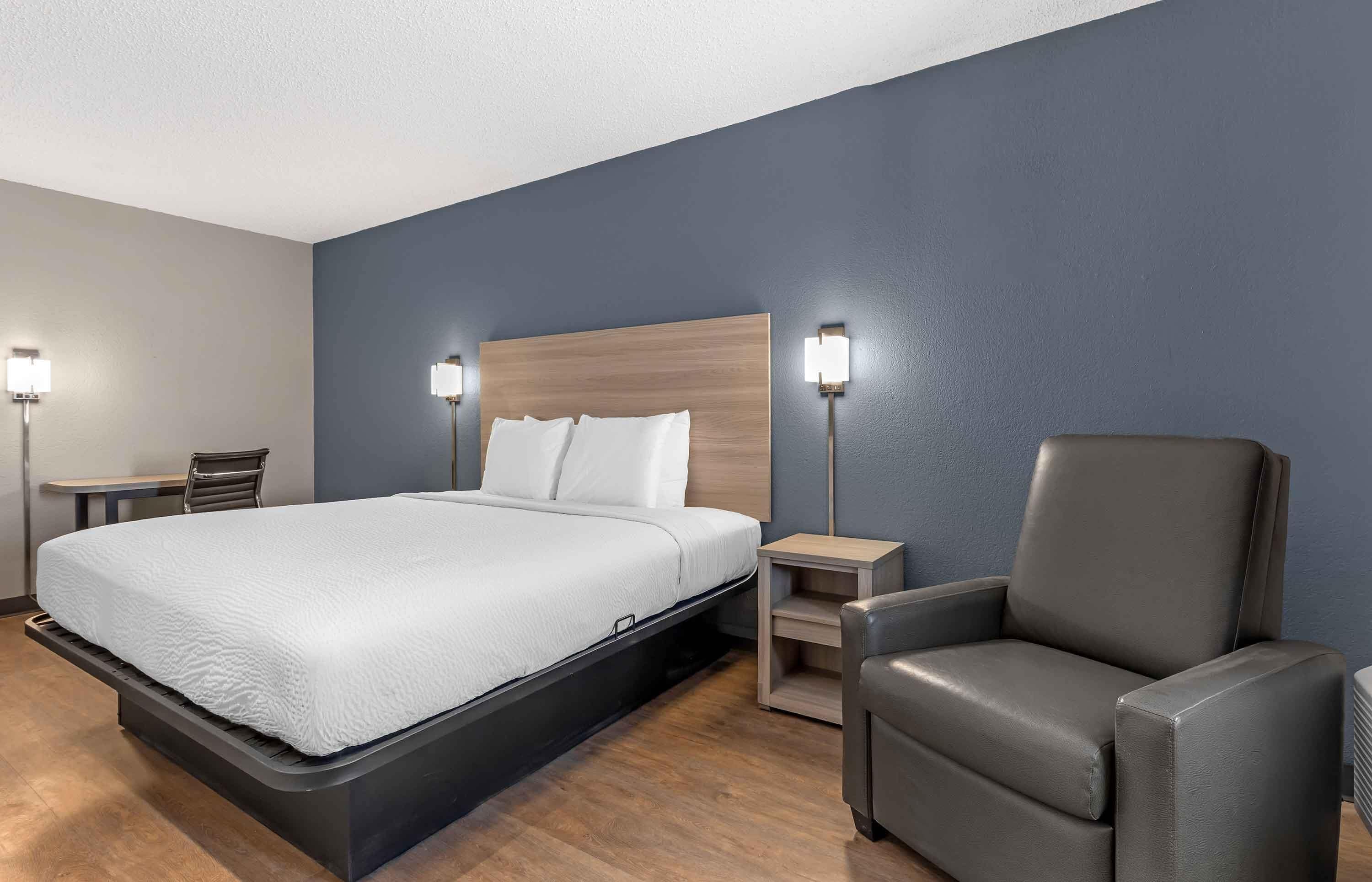 A hotel room with a bed, a chair, a nightstand, and a lamp.