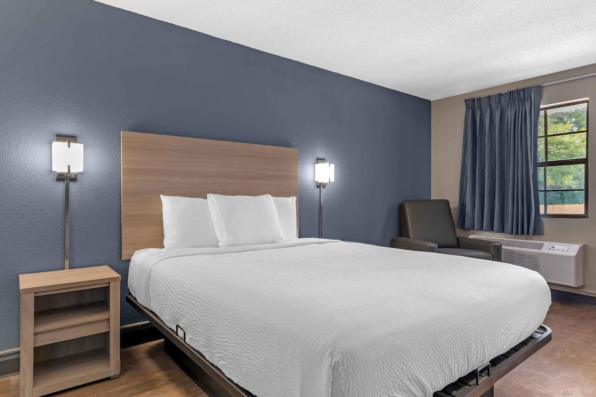 A neatly made bed with white linens, a wooden headboard, two bedside lamps, a chair, and a window with curtains.