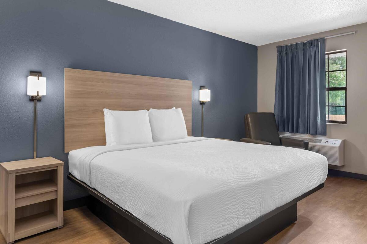 A hotel room with a large bed, two bedside tables, a chair, an air conditioning unit, and a window with curtains.