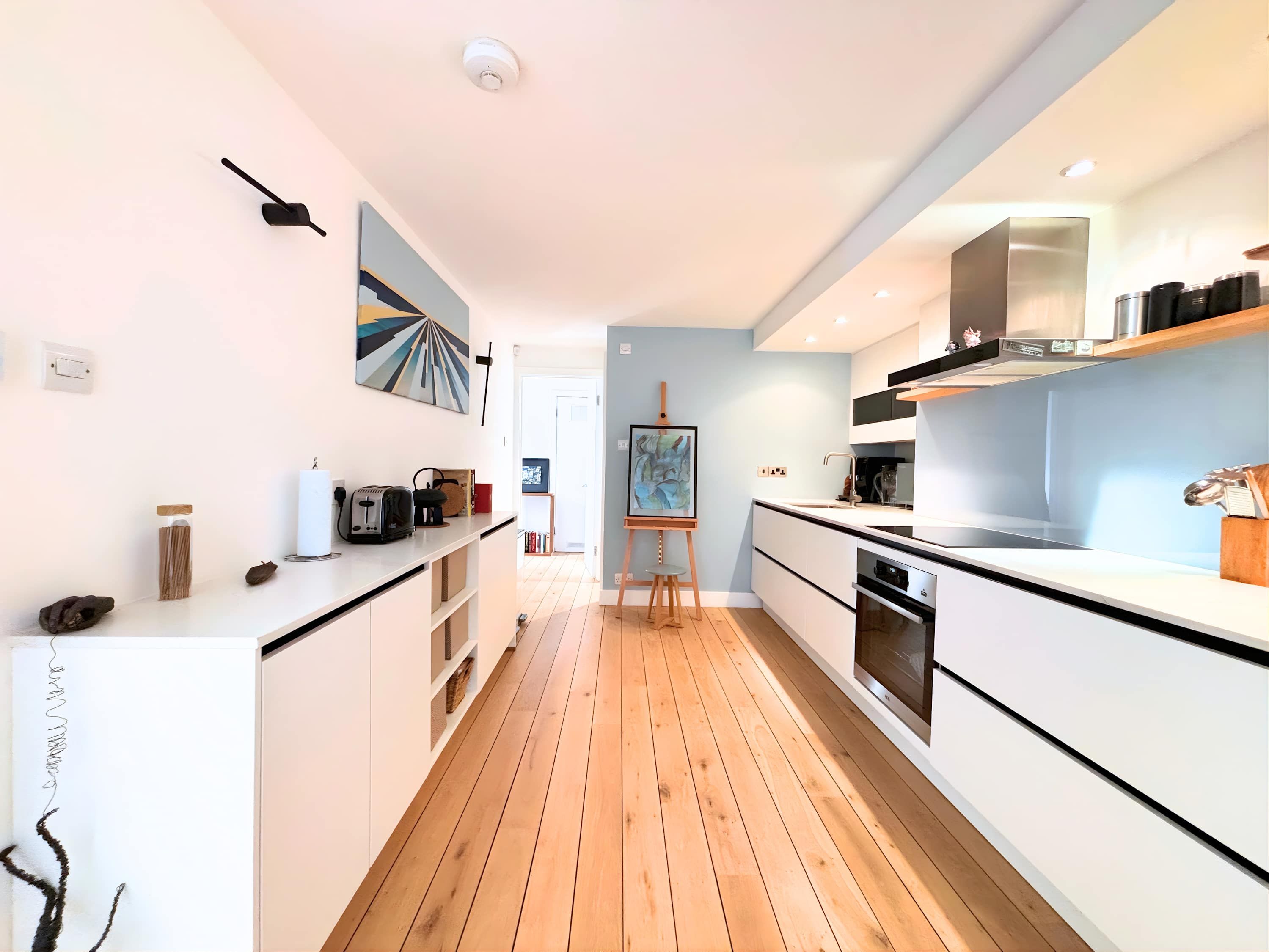 A modern kitchen with white cabinetry, a wooden floor, and a painting on the wall.