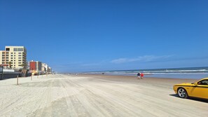 Una playa con un sendero de arena, edificios al fondo y un coche amarillo estacionado a la derecha.