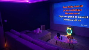 A dark room with purple lighting, a projection screen displaying text, a sofa, a coffee table with a speaker, and a small table with a vase and flowers. - Suite, SPA Jacuzzi, Cinema & Karaoke - Romantic getaway or with friends! (Saint-Joseph)