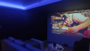 A dark room with a large screen displaying a movie poster, a small round table with remotes, and a row of sofas. - Suite, SPA Jacuzzi, Cinema & Karaoke - Romantic getaway or with friends! (Saint-Joseph)