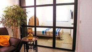 Ein Zimmer mit einer großen Schiebeglaswand, die auf einen Balkon führt, einem Sofa, einem kleinen Tisch mit einer Vase und einem Bett mit einer gestreiften Decke.