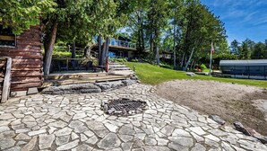 Una cabaña rústica con un patio de piedra, una fogata y una terraza de madera con vista a un área verde.