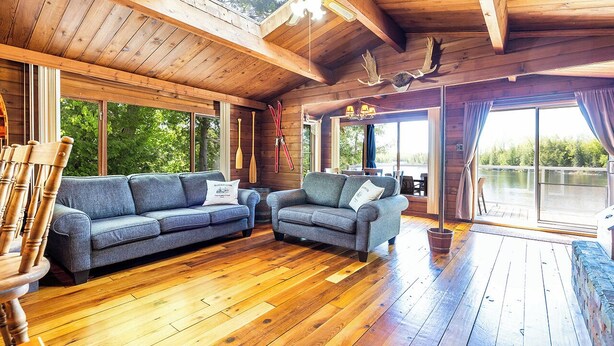 Un interior de madera con dos sofás grises, suelo de madera y ventanas amplias que ofrecen vistas a zonas verdes.