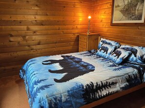 A wooden cabin bedroom with a bed featuring a bear and forest design, a nightstand, and a framed picture on the wall. - THE BUCKHORN LAKEHOUSE Peaceful Waterfront Retreat (Lakehurst)