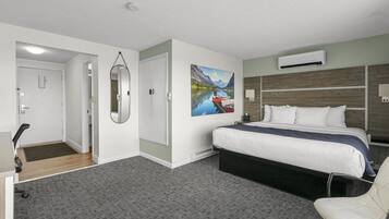 A hotel room with a bed, a desk, a chair, a painting, and an air conditioning unit.
