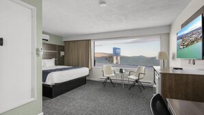 Une chambre d’hôtel équipée d’un lit, d’un bureau, d’une chaise, d’une télévision et d’une grande fenêtre donnant sur la ville et les montagnes.