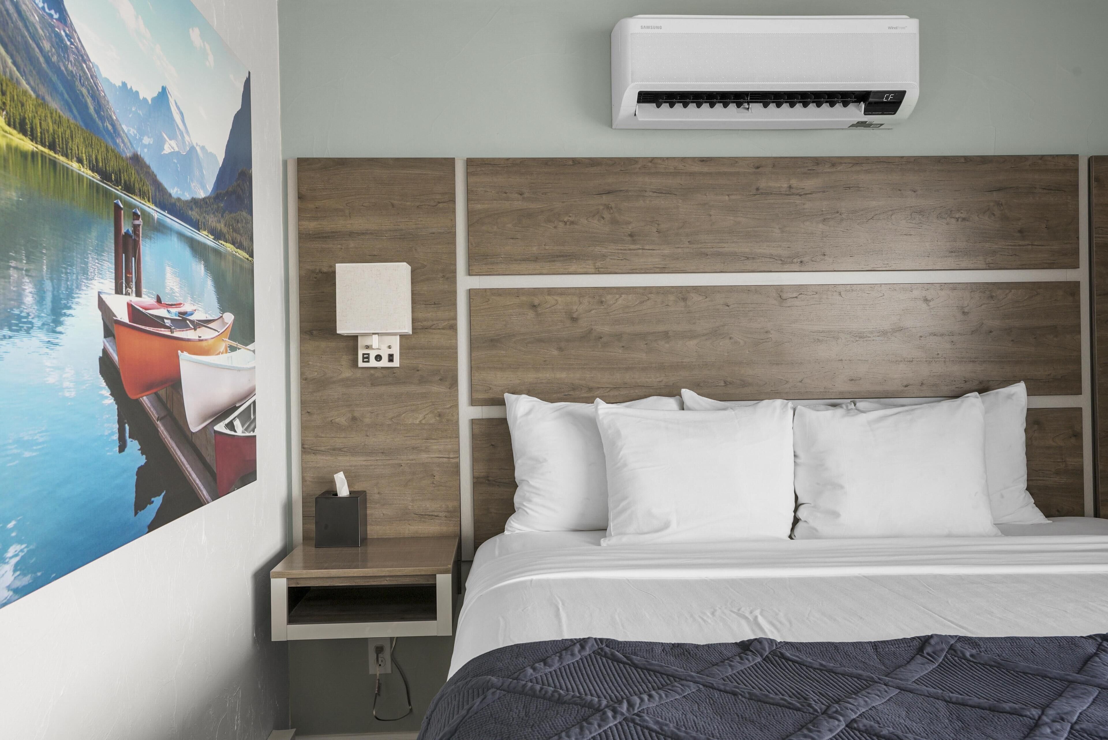 A hotel room with a bed, white pillows, a nightstand with a lamp, an air conditioner, and a painting of a lake with boats.