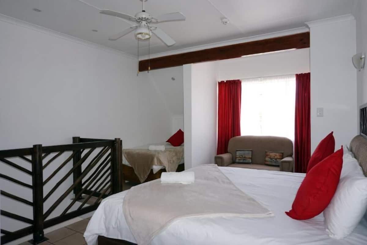 Habitación de hotel con cama, una silla, ventana con cortinas rojas, ventilador de techo y viga de madera.