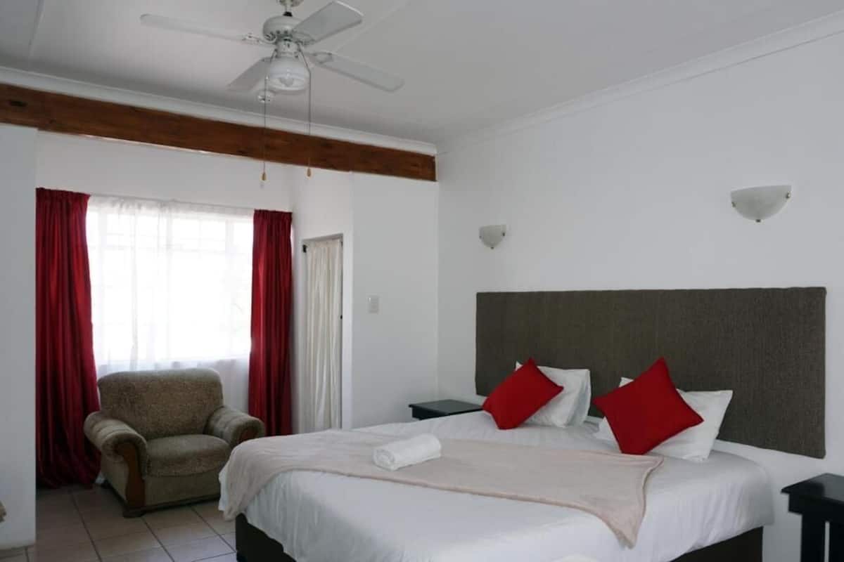 Un dormitorio con cama, una silla, una mesita de noche, un ventilador de techo y una ventana con cortinas.
