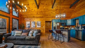 Ein gemütliches Blockhaus-Interieur mit Holzbalkendecke, einem Wohnbereich mit Sofa und Sesseln, einer Küche mit blauen Schränken und Kunstwerken an den Wänden.