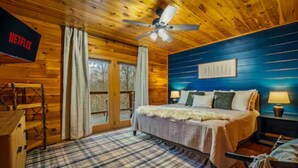 Ein gemütliches Zimmer mit einem Bett, einem Deckenventilator, einem Fernseher mit 'NETFLIX' und einem großen Fenster mit Vorhängen.