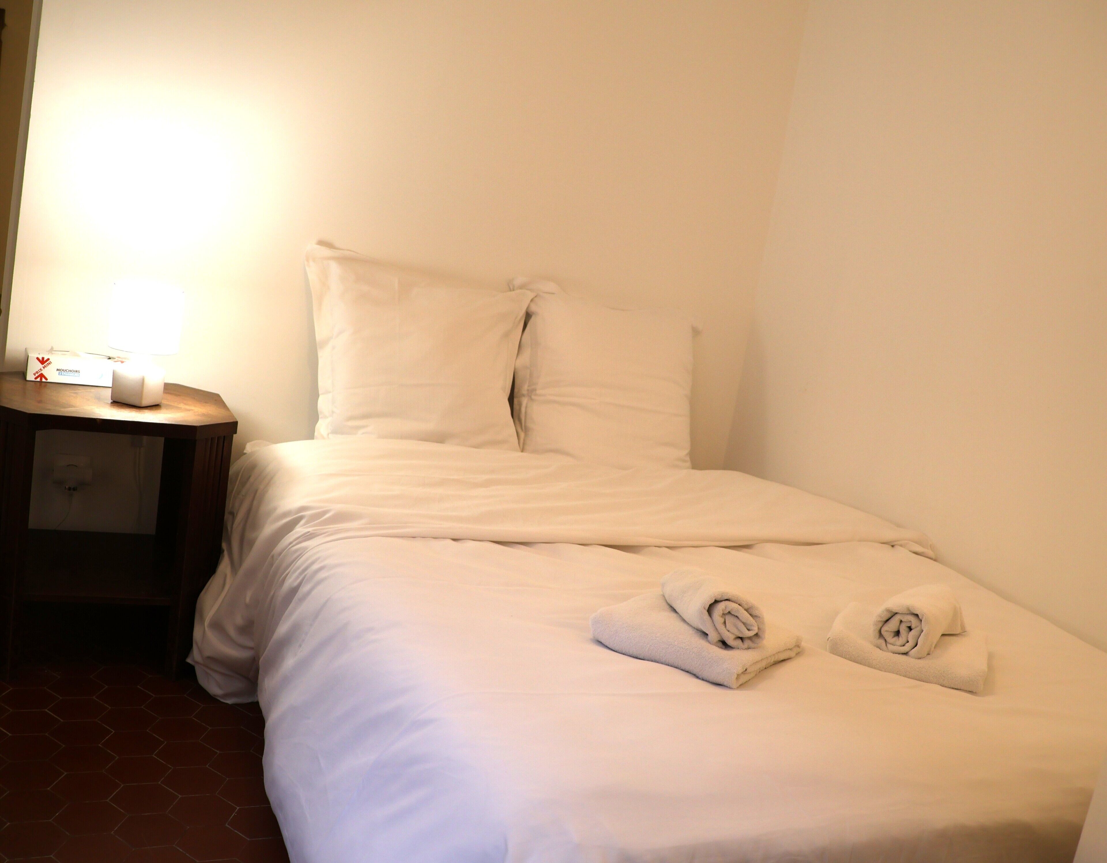 Una cama bien hecha con sábanas blancas y dos toallas enrolladas, una mesita de noche con una lámpara y una caja, y un piso de baldosas.