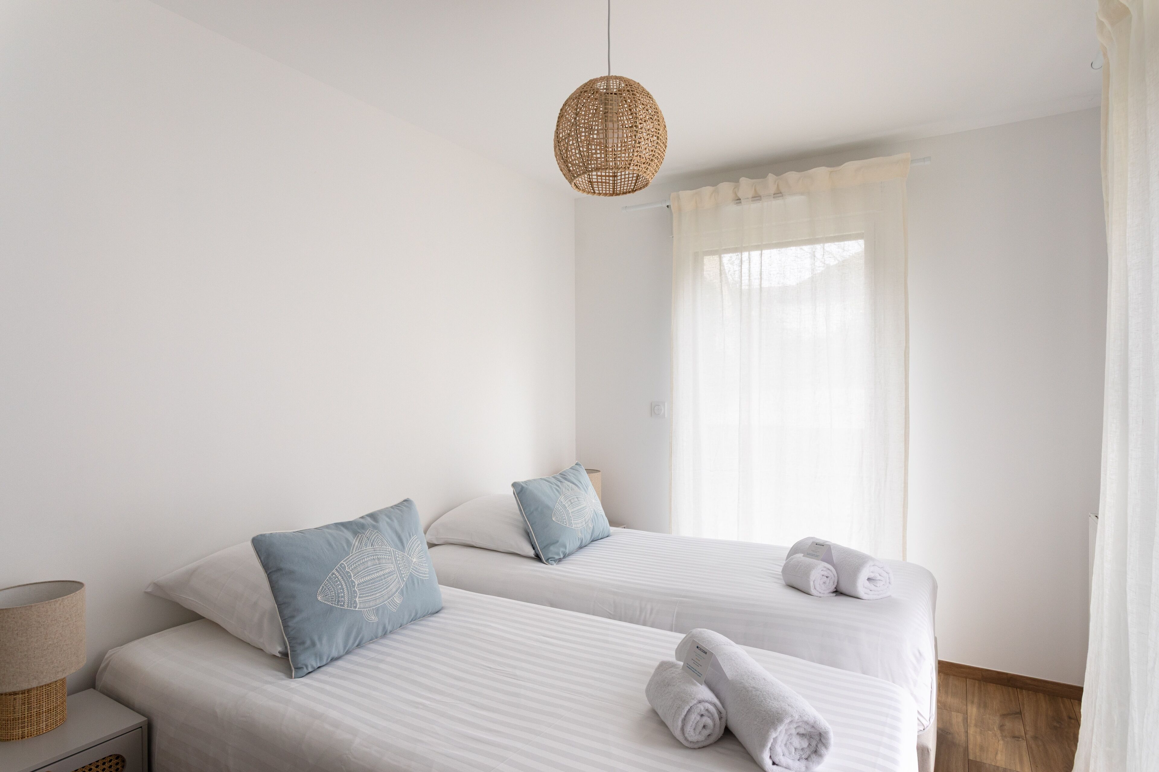 A hotel room with two beds, a hanging rattan light fixture, a bedside table with a lamp, and a window with sheer curtains.