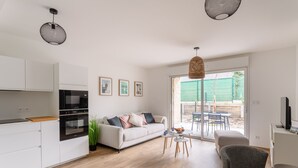 Una cocina moderna con armarios blancos y un horno negro, un sofá blanco con cojines, una mesa de comedor con sillas y un amplio ventanal corredero al exterior.