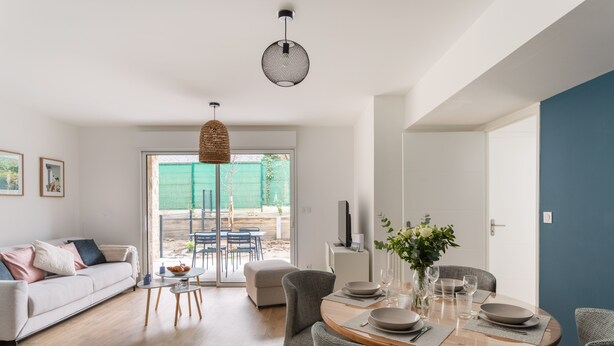 Una sala de estar moderna con comedor, que incluye un sofá, sillas, una mesa de comedor preparada para cuatro personas y un amplio ventanal con vistas a un jardín.