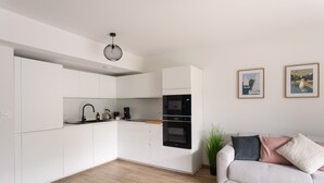 Una cocina moderna con armarios blancos, un horno negro y una encimera de madera. Hay un sofá blanco con cojines y una planta en la zona de estar.