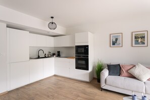 Une cuisine moderne avec des meubles blancs, un four noir et un plan de travail en bois. Le salon est aménagé avec un canapé blanc agrémenté de coussins et d’une plante.