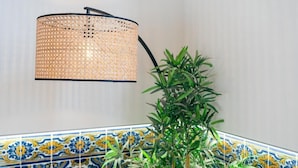 Una lampada con paralume intrecciato e base nera, una pianta in vaso e una parete piastrellata con motivi blu e gialli.