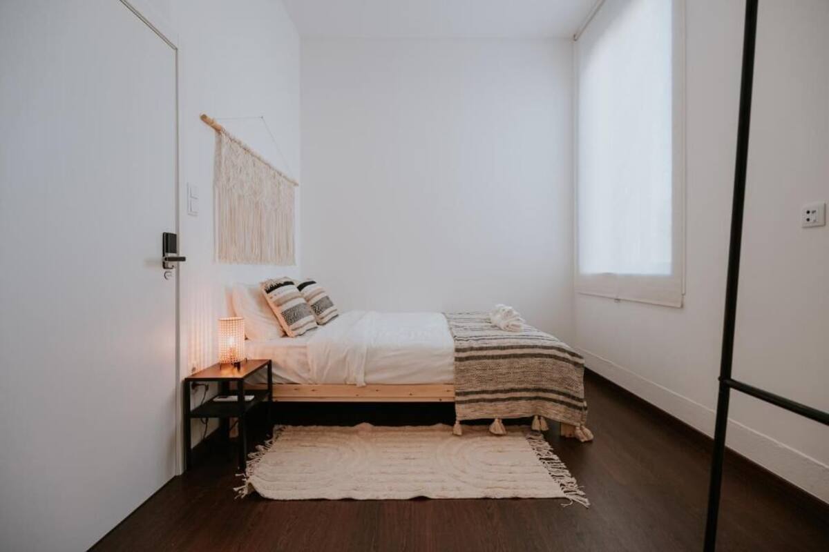 A small, minimalist bedroom with a single bed, a bedside table with a lamp, a macrame wall hanging, and a window providing natural light.