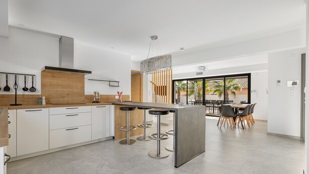 Una cocina moderna con armarios blancos, encimera de madera y una zona de bar con taburetes. Se ve un comedor con una mesa y sillas a través de una puerta de vidrio.