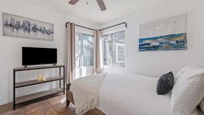A bedroom with a bed, a TV on a stand, a ceiling fan, and two windows with curtains. - Pacific Beach Bungalow - On Boardwalk (San Diego)