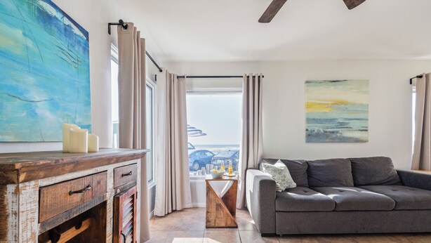 A living room with a grey sofa, a wooden sideboard with drawers, a large abstract painting, and a view of parked cars through the window. - Pacific Beach Bungalow - On Boardwalk (San Diego)