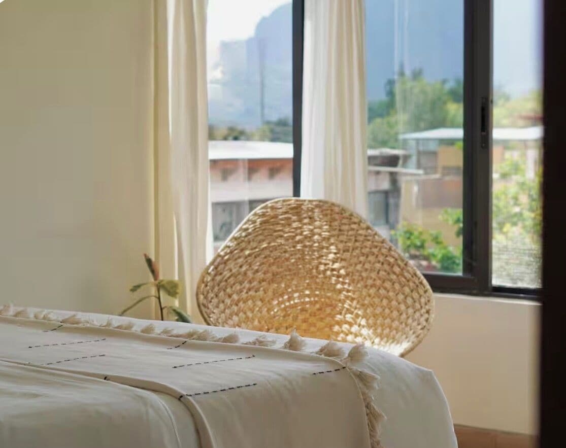 Una cama con cubrecama blanco, una silla de mimbre y vistas a edificios a través de una ventana.