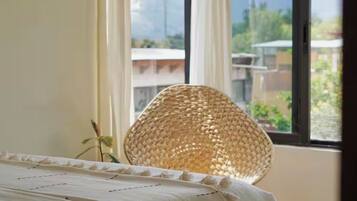 Una cama con cubrecama blanco, una silla de mimbre y vistas a edificios a través de una ventana.