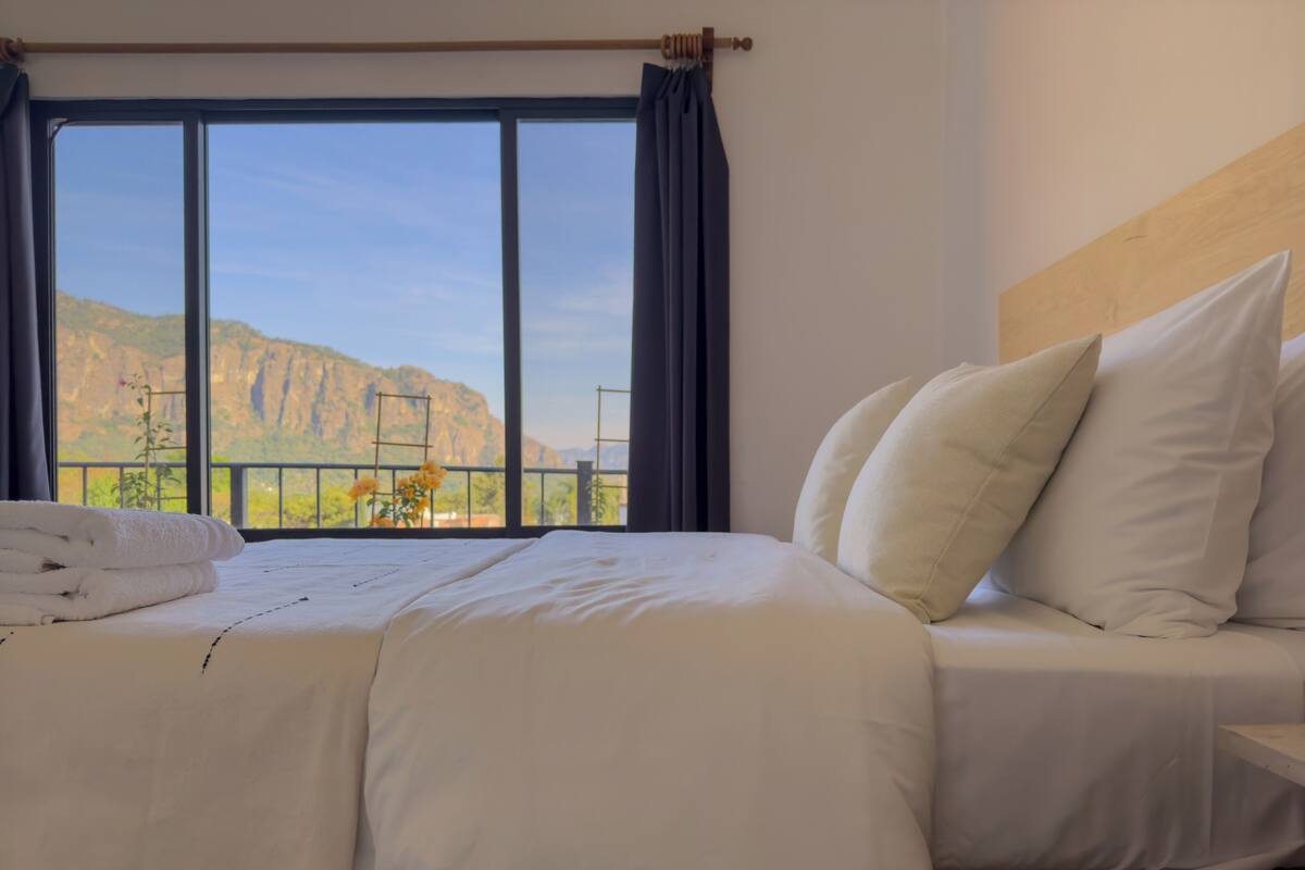 A hotel room with a large window, a neatly made bed with white linens, and a view of mountains.