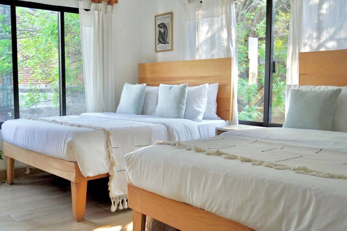 A bedroom with two beds, a wooden headboard, white bedding, and a large window with a view of greenery.
