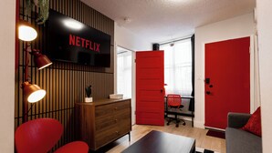 Ein modernes Wohnzimmer mit Flachbildfernseher, einer roten Tür, einem hölzernen Schminktisch, einem roten Sessel und einem grauen Sofa.