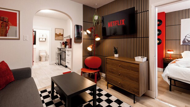 Ein modernes Hotelzimmer mit einem Flachbildfernseher, einer Holzkombination, einem roten Sessel, einem kleinen Tisch und einem karierten Teppich.