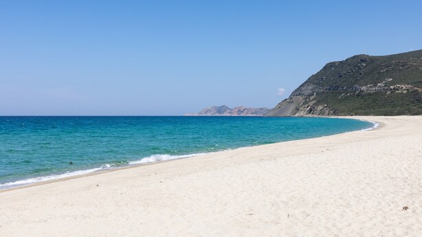 A beach with clear blue water, a sandy shore, and a mountain in the background.
