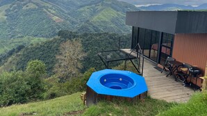 Un bain à remous sur une terrasse en bois, avec vue sur des collines verdoyantes et des montagnes.