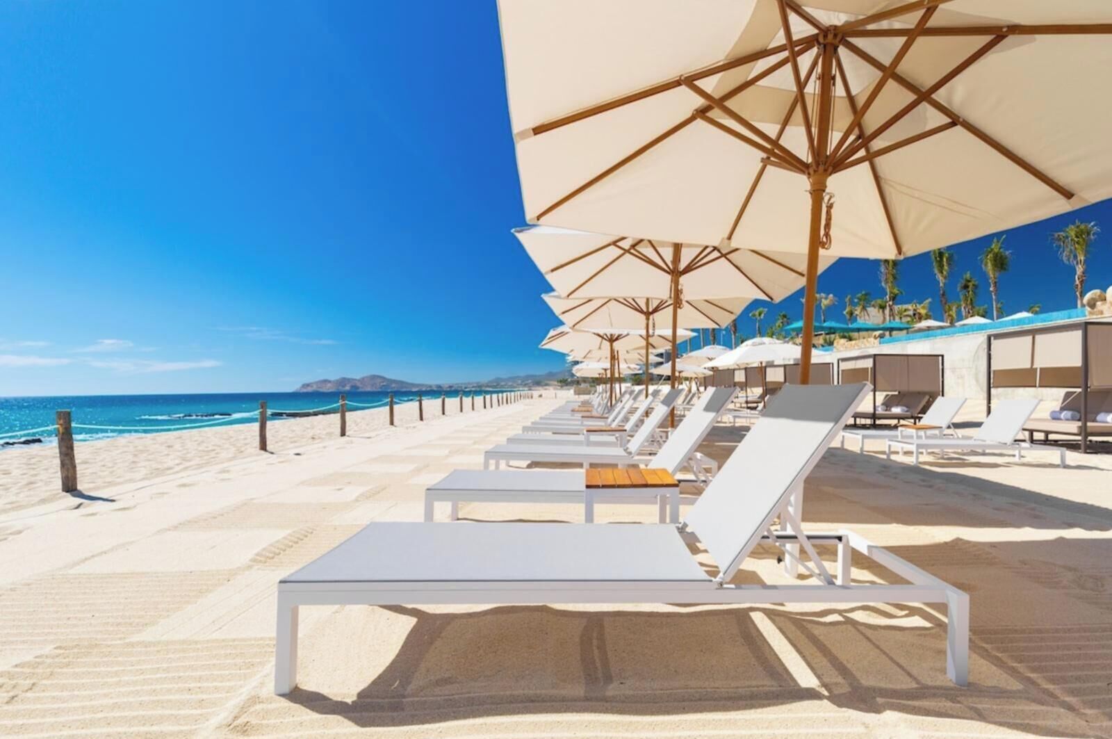 A beachside hotel with rows of white sun loungers under large umbrellas, a sandy beach, and a clear blue sky.