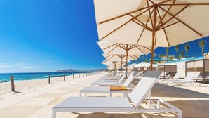 Ein Strandhotel mit Reihen aus weißen Liegestühlen unter großen Sonnenschirmen, ein Sandstrand und ein klarer, blauer Himmel.