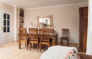 A dining area with a wooden table and chairs, a mirror with an ornate frame, and a bookshelf with books and decorative items.